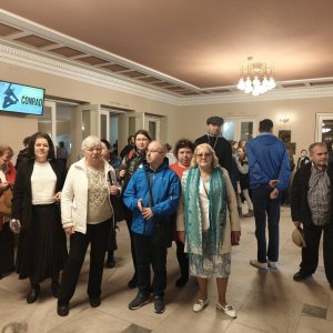 Pamiątkowe zdjęcie grupowe uczestników i opiekunów Środowiskowego Domu Samopomocy w Swarzędzu stojących w jasnym teatralnym foyer pod wiszącym żyrandolem. Na ścianie po lewej stronie widoczny jest włączony ekran informacyjny z plakatem i napisem „CONRAD”. Grupa w jesiennych kurtkach, płaszczach i szalach pozuje przed wejściem na widownię spektaklu baletowego. Wyjście odbyło się w ramach projektu „Teatr bez barier”, współfinansowanego ze środków Samorządu Województwa Wielkopolskiego.