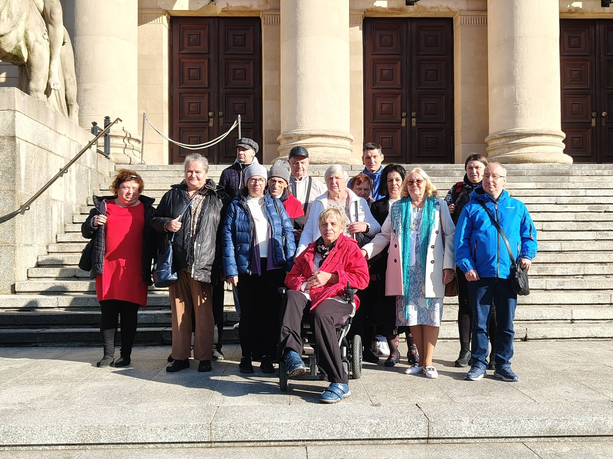 Pamiątkowe zdjęcie grupowe uczestników i opiekunów Środowiskowego Domu Samopomocy w Swarzędzu na schodach przed wejściem do Teatru Wielkiego im. Stanisława Moniuszki w Poznaniu, w pełnym słońcu. W centralnej części kadru siedzi uśmiechnięta kobieta na wózku inwalidzkim w czerwonej kurtce, otoczona przez grupę stojącą na kamiennych stopniach. W tle widoczne są masywne kolumny neoklasycystycznego portyku, drewniane wrota teatru oraz fragment rzeźby lwa. Zdjęcie wykonano podczas wyjścia na spektakl baletowy „Conrad” zrealizowanego w ramach projektu „Teatr bez barier”, współfinansowanego ze środków Samorządu Województwa Wielkopolskiego.