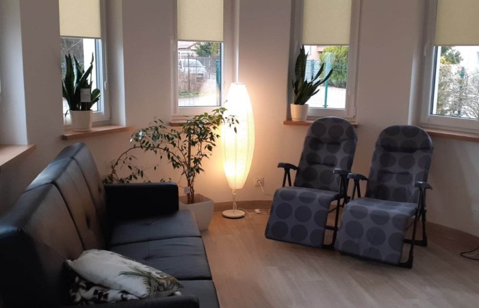 Zdjęcie przedstawia jasny pokój z wieloma oknami, w którym znajdują się sofa oraz dwa rozkładane krzesła w stylu leżaków. W pomieszczeniu stoi duża lampa, która rzuca ciepłe światło na cały pokój. Na parapetach widoczne są roślinki.