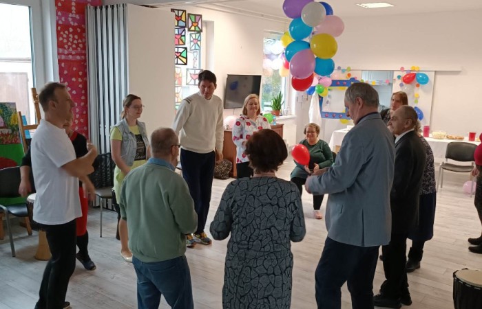 Zdjęcie przedstawia grupę osób bawiących się w kółeczku w sali, która jest wystrojona imprezowo. Widoczne są balony oraz instrumenty muzyczne, takie jak bębenki, rozstawione na podłodze.