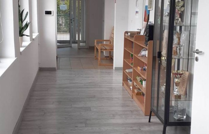 Zdjęcie przedstawia główny korytarz wewnątrz budynku, zaraz po wejściu. Na ścianach widoczne są obrazy, a także przeszklony regał z nagrodami i pucharami oraz komoda wypełniona różnymi przedmiotami.