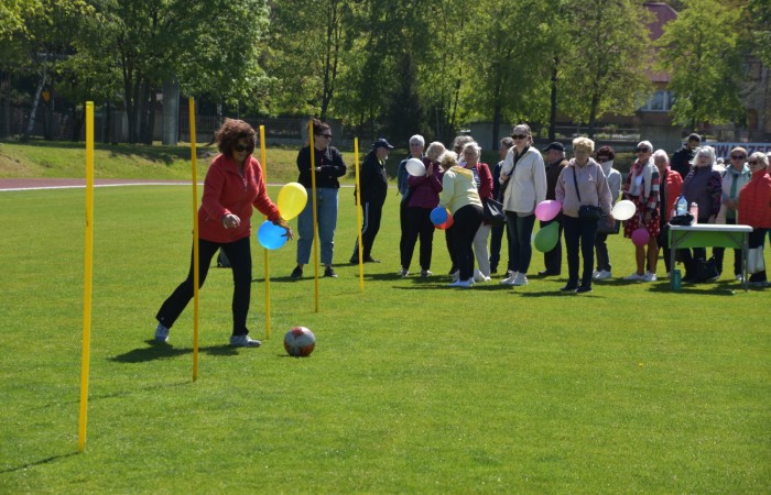 Zdjęcie przedstawia kobietę w czerwonej bluzie prowadzącą piłkę pomiędzy żółtymi tyczkami na zielonym boisku. W jednej ręce trzyma dwa balony – żółty i niebieski. W tle znajduje się grupa seniorów trzymających balony, czekających na swoją kolej do zabawy. 