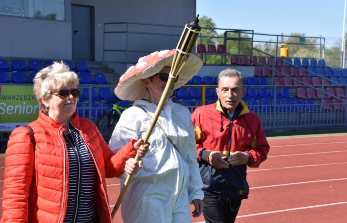 Zdjęcie przedstawia trójkę uczestników wydarzenia na stadionie. Kobieta w czerwonej kurtce trzyma drewnianą pochodnię, a obok niej stoi osoba przebrana za muchomora – w białym kombinezonie z dużym czerwonym kapeluszem w białe kropki. Obok znajduje się starszy mężczyzna w czerwono-żółtej kurtce. 