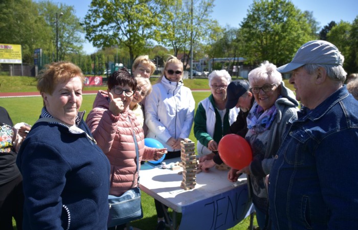 Zdjęcie przedstawia grupę seniorów grających w Jengę na świeżym powietrzu. Na stole ustawiona jest drewniana wieża, a uczestnicy z uśmiechem i entuzjazmem biorą udział w grze. Niektórzy trzymają kolorowe balony. 