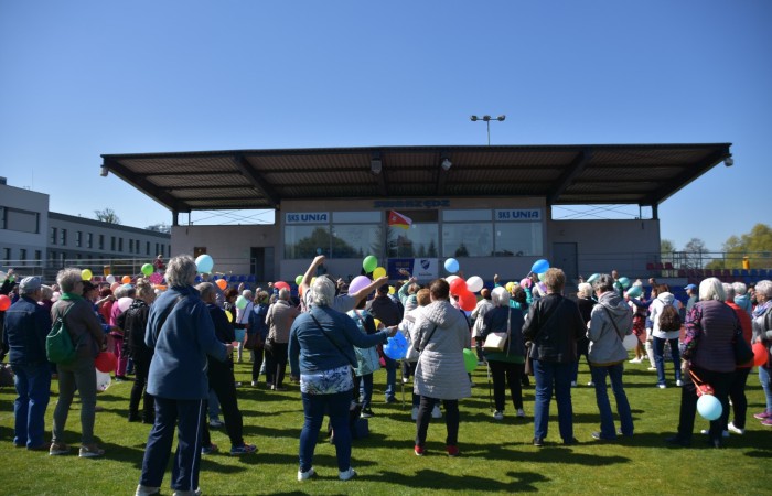 Zdjęcie przedstawia dużą grupę seniorów zgromadzonych na boisku przed trybuną stadionu. Wiele osób trzyma kolorowe balony i patrzy w stronę sceny. Na budynku trybuny widnieją napisy „SKS UNIA” oraz „Swarzędz”. 