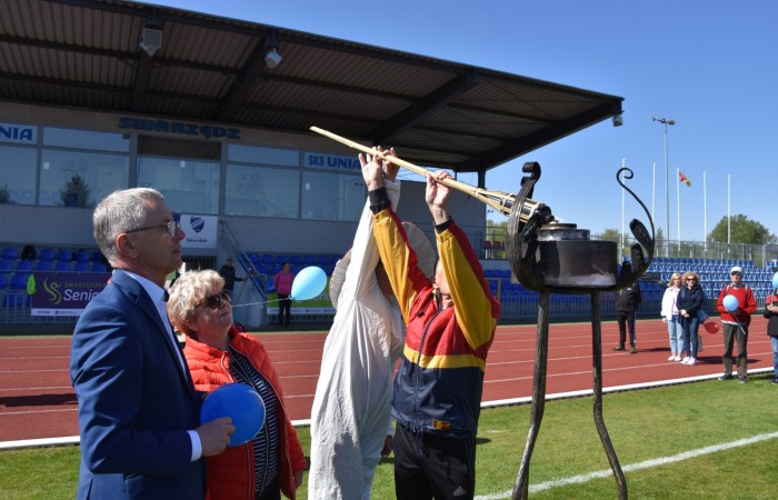 Zdjęcie przedstawia mężczyznę w czerwono-żółtej kurtce podnoszącego pochodnię do metalowego kociołka na wysokim stojaku. Obok niego stoi elegancko ubrany mężczyzna w garniturze oraz kobieta w czerwonej kurtce. W tle widoczna jest trybuna stadionu i kilka osób trzymających balony. 