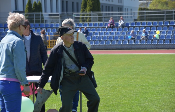 Zdjęcie przedstawia starszego mężczyznę w czapce i czarnej kurtce biorącego udział w zawodach. Mężczyzna jest w ruchu, trzymając duży, szary przedmiot przypominający but lub worek. Wokół niego stoją inni uczestnicy, a w tle widoczna jest trybuna z niebieskimi krzesłami. 