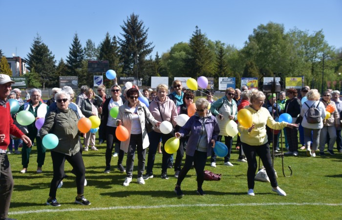 Zdjęcie przedstawia grupę seniorów na boisku uczestniczących w zabawie z balonami. Niektóre osoby unoszą ręce, inni trzymają balony w różnych kolorach – żółtym, pomarańczowym, zielonym i niebieskim. Tło stanowi boisko otoczone drzewami oraz reklamy na ogrodzeniu.