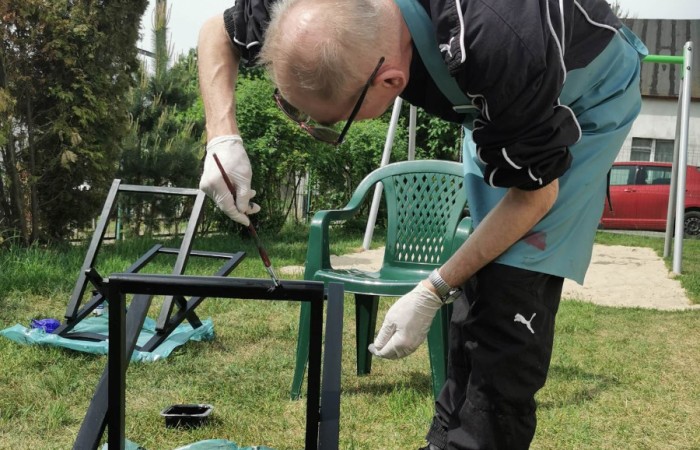 Zdjęcie przedstawia mężczyznę malującego metalowy stelaż krzesła na czarno. Jest pochylony nad krzesłem, trzyma pędzel i maluje konstrukcję. Ma na sobie czarną sportową kurtkę, niebieski fartuch ochronny i rękawiczki. Tło stanowi ogród z zieloną trawą, krzesłem ogrodowym i czerwonym samochodem zaparkowanym w oddali. 