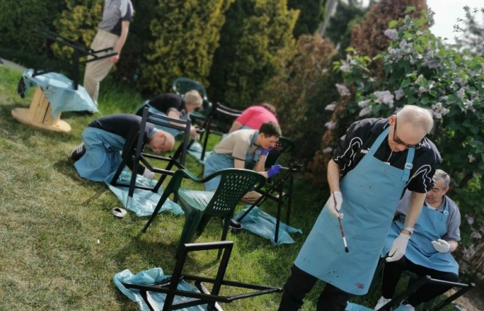 Zdjęcie przedstawia grupę osób malujących metalowe krzesła w ogrodzie. Każdy uczestnik nosi jasnoniebieski fartuch ochronny i rękawiczki. Część osób pracuje w pozycji pochylonej, inne siedzą na plastikowych krzesłach. W tle widoczna jest roślinność, drzewa oraz budynek.