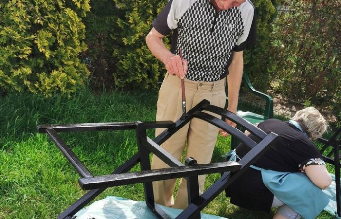 Zdjęcie przedstawia starszego mężczyznę malującego metalowy stelaż krzesła. Stoi przy drewnianym stole, na którym umieszczony jest stelaż, i dokładnie nakłada czarną farbę. Ma na sobie koszulkę polo w czarno-biały wzór oraz jasne spodnie. W tle znajduje się ogród z zielonymi krzewami.