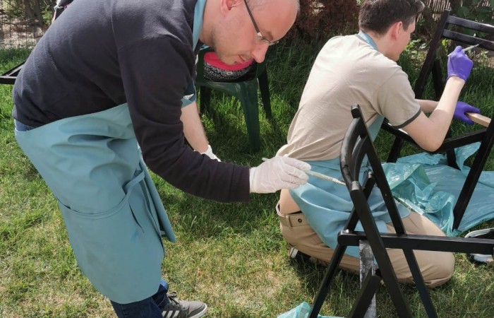 Zdjęcie przedstawia mężczyznę w okularach koncentrującego się na malowaniu krzesła. Jest pochylony, trzyma pędzel i uważnie nakłada czarną farbę na metalową konstrukcję. W tle widać innych uczestników warsztatów malujących swoje krzesła, a także ogród z zielenią i fioletowymi kwiatami.