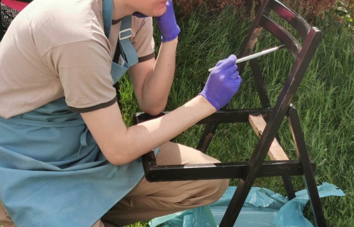 Zdjęcie przedstawia mężczyznę kucającego przy krześle i malującego jego stelaż. Jest ubrana w niebieski fartuch ochronny, beżową koszulkę i spodnie. W rękach trzyma pędzel i kuwetę z czarną farbą. Pracuje na zielonej trawie, a w tle widoczne są krzewy i płot.