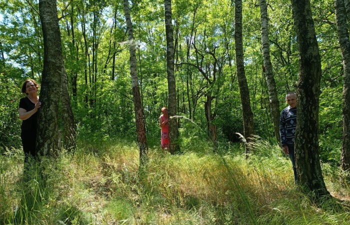 Zdjęcie przedstawia troje ludzi stojących między drzewami w lesie. Każda z osób wygląda zza drzewa, uśmiechając się. Po lewej stronie kobieta w czarnej sukience, na środku kobieta w różowej sukience, a po prawej stronie mężczyzna w granatowej bluzie. Las jest pełen wysokich, smukłych drzew i soczystej zieleni.