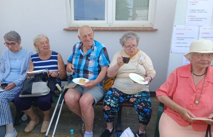 Sześć starszych osób siedzi na ławce na zewnątrz budynku, spożywając ciasto i napoje. Są ubrane w letnie stroje, jedna z kobiet ma laskę. Za nimi znajduje się plansza z harmonogramem wydarzenia. 