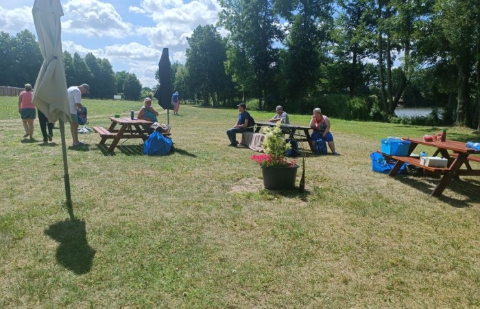 Grupa osób siedzi przy drewnianych ławkach ustawionych na trawiastej plaży 
