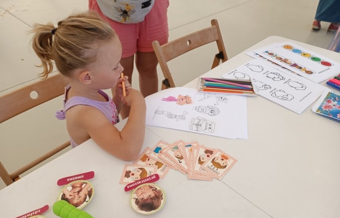 Dziewczynka w jasnofioletowej koszulce siedzi przy stole i trzyma kredkę przy ustach, patrząc na kartki z ilustracjami do pokolorowania. Na stole leżą karty edukacyjne z emocjami, kredki i inne materiały plastyczne.