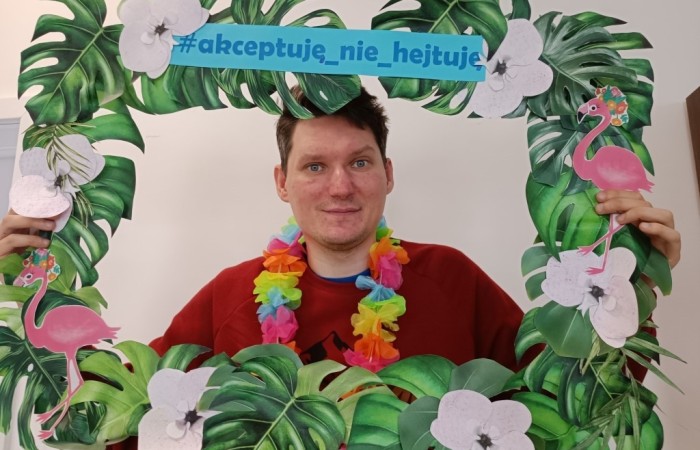 Młody mężczyzna w czerwonej bluzie trzyma ozdobną ramkę z tropikalnym motywem i napisem #akceptuję_nie_hejtuję. Ma na szyi kolorowy naszyjnik z kwiatów i patrzy w obiektyw z lekkim uśmiechem.