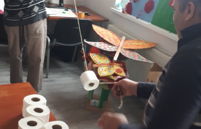 Na zdjęciu dwie osoby biorą udział w zabawie, trzymając sznurek, który przepleciony jest przez rolkę papieru toaletowego. Na stole obok nich leżą dodatkowe rolki papieru. W tle widoczne są rękodzieła w kształcie kwiatów i motyli.