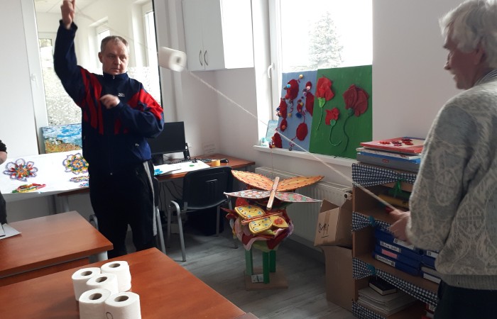 Zdjęcie przedstawia dwie osoby uczestniczące w grze lub zabawie, trzymające sznurek przepleciony przez rolkę papieru toaletowego. Na stole obok nich widoczne są kolejne rolki papieru. W pokoju znajdują się różne rękodzieła w kształcie kwiatów oraz motyli.