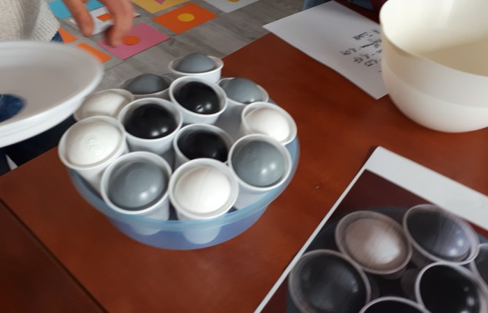 Zdjęcie przedstawia miskę z plastikowymi kubkami, w których znajdują się piłki w kolorach szarym, białym i czarnym.