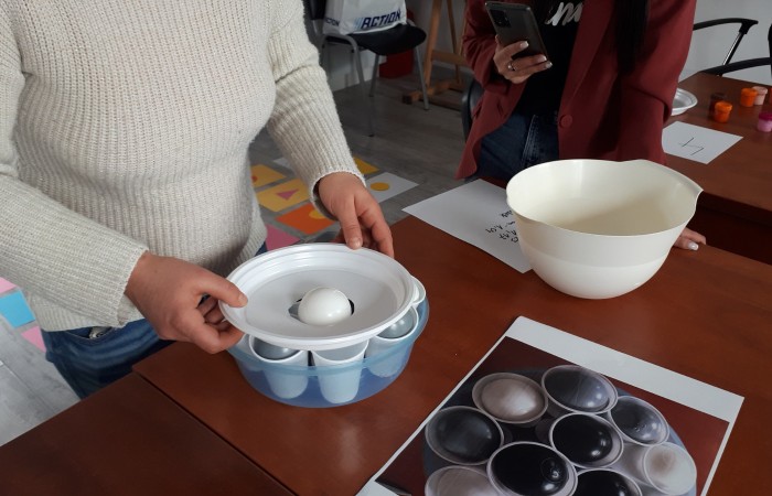 Zdjęcie przedstawia osobę trzymającą plastikowy talerzyk z wyciętą dziurą pośrodku nad miską z kubkami, w których znajdują się piłki. Osoba wydaje się wrzucać białą piłkę przez dziurę w talerzyku do kubków poniżej.