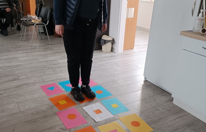Zdjęcie przedstawia osobę uczestniczącą w zabawie, stojącą na kolorowej kartce papieru. Dookoła niej na podłodze rozłożone są kartki papieru z kształtami w różnych kolorach.
