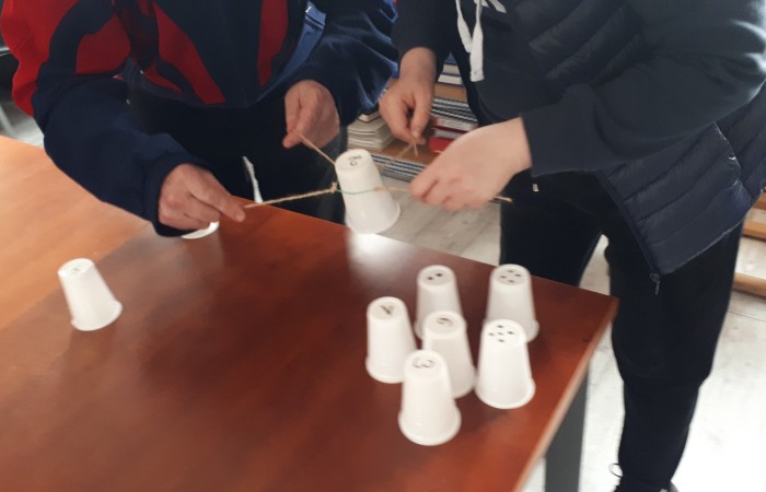 Zdjęcie przedstawia dwie osoby uczestniczące w grze lub zabawie, trzymające końcówki sznurka, który jest zawinięty wokół plastikowego kubka. Na stole widoczne są pozostałe kubki, ustawione do góry dnem. Kubki mają na spodach numery oraz inne wzory.