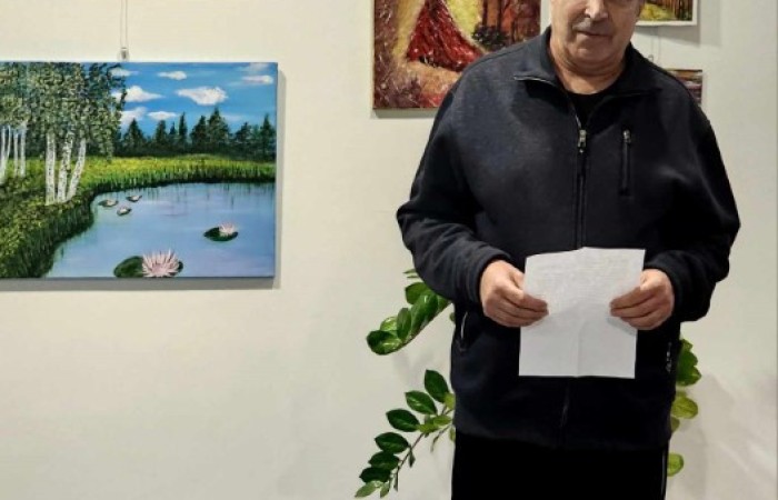 starszy mężczyzna w czarnej bluzie trzyma kartkę papieru, stojąc przed białą ścianą, na której zawieszono kilka obrazów przedstawiających pejzaże, postaci i abstrakcyjne kompozycje.