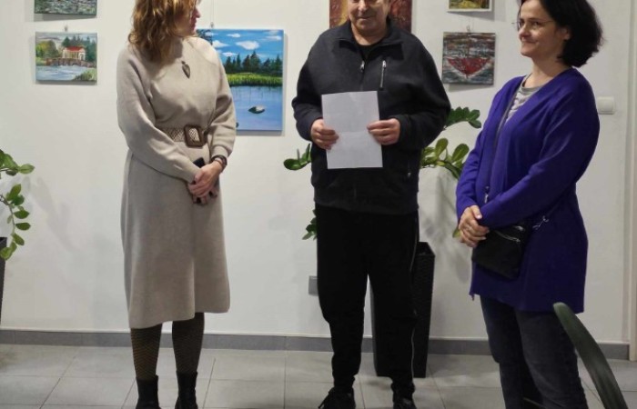 Mężczyzna i dwie kobiety na tle wystawy scena rozmowy podczas wernisażu, trzy osoby stoją na tle białej ściany z pejzażami i portretami.