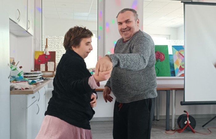 Mężczyzna w szarym swetrze i kobieta w czarnej bluzie oraz różowej sukience tańczą razem, trzymając się za ręce. Uśmiechają się i patrzą na siebie. W tle widać białą kuchnię z blatami pełnymi różnych przedmiotów oraz kolorowe obrazy na sztalugach. Na suficie widoczne są wielokolorowe światła dekoracyjne.