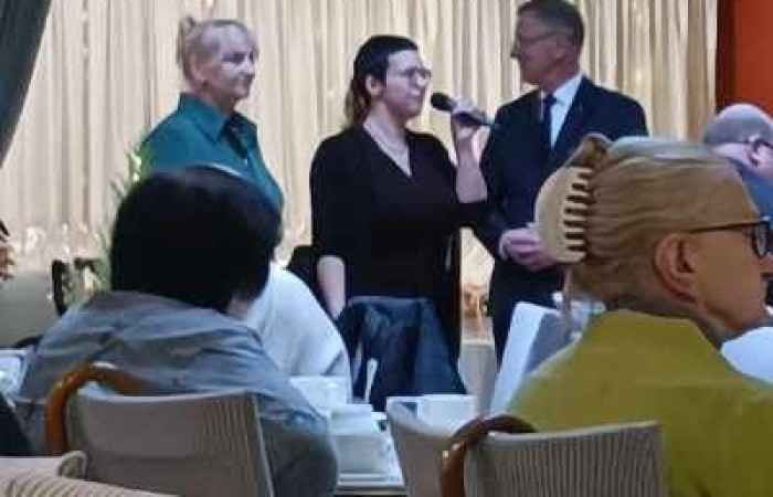 trzy osoby stoją i rozmawiają przy mikrofonie w eleganckim wnętrzu z jasnymi zasłonami. Widzowie siedzą przy stolikach w tle. 