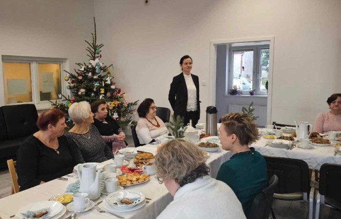 kobieta w czarnej marynarce stoi i przemawia do grupy osób siedzących przy stole. W tle znajduje się udekorowana choinka oraz okna z widokiem na zewnątrz. 