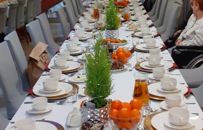 długi stół w jasnym wnętrzu, nakryty białym obrusem, udekorowany zielonymi gałązkami i pomarańczami. Nakrycia składają się z białej porcelany, złotych podkładek i sztućców. W tle schody z czerwonymi gwiazdami i świąteczne oświetlenie.