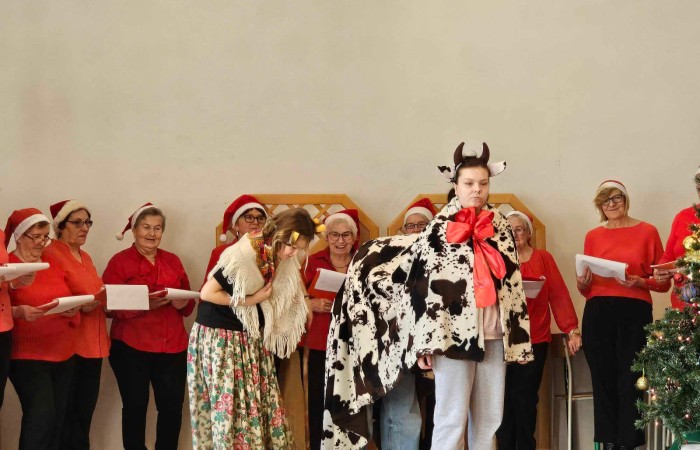Kolędnicy na scenie a przed nimi scenka odgrywana, osoba przebrana za kobietę i dwie osoby za krowę