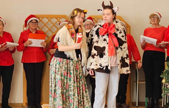 Kolędnicy na scenie a przed nimi scenka odgrywana, osoba przebrana za kobietę i dwie osoby za krowę
