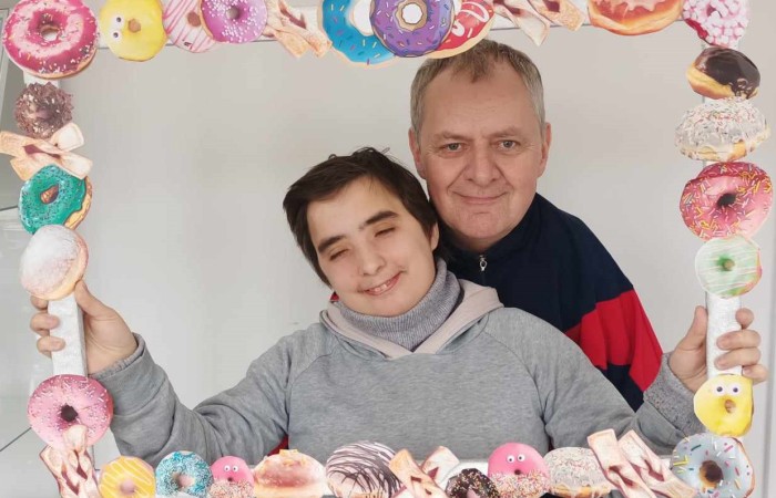 Dwie osoby pozują do zdjęcia, trzymając ozdobną ramkę. Mężczyzna w czerwono-niebieskiej bluzie stoi za młodą osobą w szarej bluzie z kapturem, która uśmiecha się delikatnie. Ramka ozdobiona jest grafikami słodkich wypieków. 