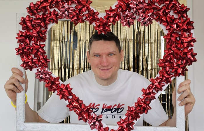 Mężczyzna w białej koszulce z napisem „Las Vegas” trzyma tę samą dekoracyjną ramkę w kształcie serca, uśmiechając się do aparatu. Tło również stanowią złote, połyskujące zasłony.