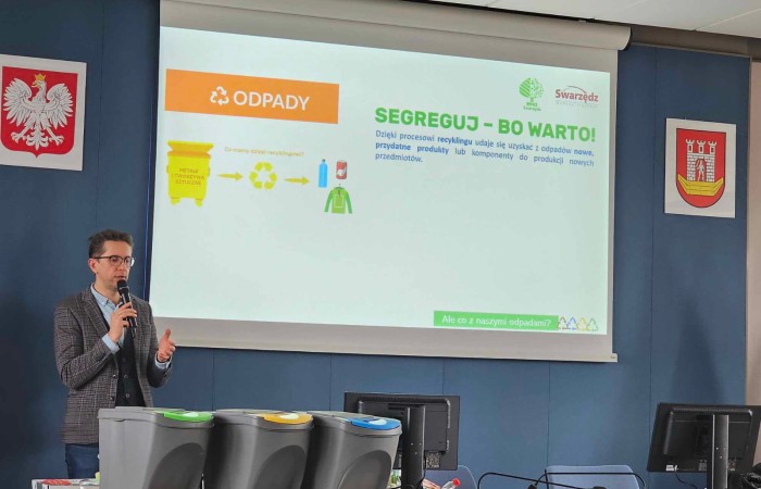 Mężczyzna w garniturze z mikrofonem stoi przed ekranem, na którym wyświetlana jest prezentacja na temat segregacji odpadów. Na stole znajdują się trzy pojemniki do segregacji śmieci. Na ekranie widoczny jest napis „Segreguj – bo warto!”.