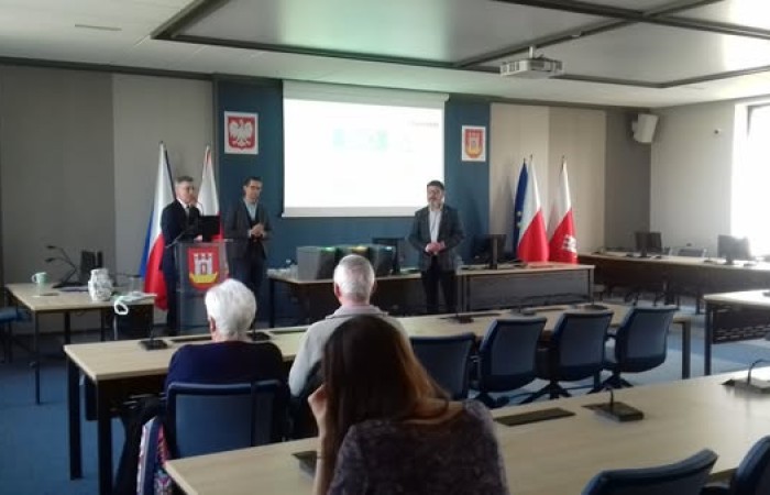 W dużej sali konferencyjnej trzech mężczyzn stoi przed publicznością, prowadząc prezentację na temat segregacji odpadów. W tle widać flagi Polski i Unii Europejskiej oraz godło miasta.