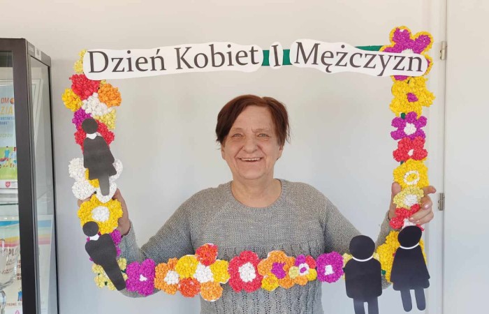 Starsza kobieta w szarym swetrze trzyma dekoracyjną ramkę z napisem „Dzień Kobiet i Mężczyzn” ozdobioną kolorowymi kwiatami z bibuły i sylwetkami postaci. Kobieta uśmiecha się do aparatu. W tle biała ściana i witryna z pucharami.