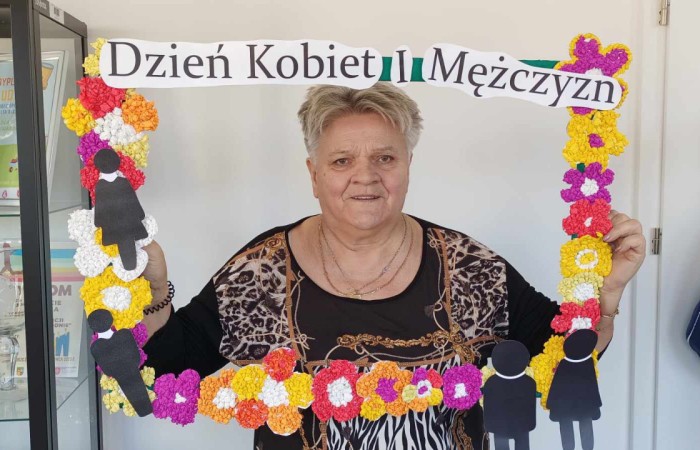 Starsza kobieta w bluzce w panterkę trzyma dekoracyjną ramkę z napisem „Dzień Kobiet i Mężczyzn”. Kobieta uśmiecha się do aparatu, w tle znajduje się przeszklona szafa z nagrodami i pucharami.