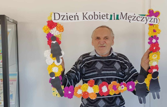 Starszy mężczyzna z siwą brodą i swetrem w geometryczne wzory trzyma dekoracyjną ramkę z napisem „Dzień Kobiet i Mężczyzn”. W tle przeszklona szafa z nagrodami. 