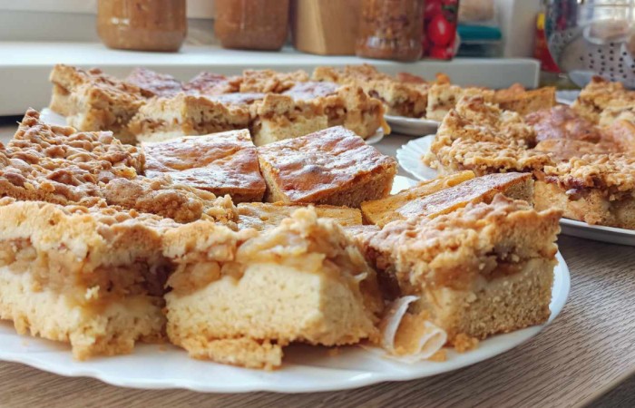 Talerze z pokrojonym ciastem jabłkowym o złocistej kruszonce stoją na drewnianym blacie. W tle słoiki z przyprawami oraz kuchenne akcesoria. 