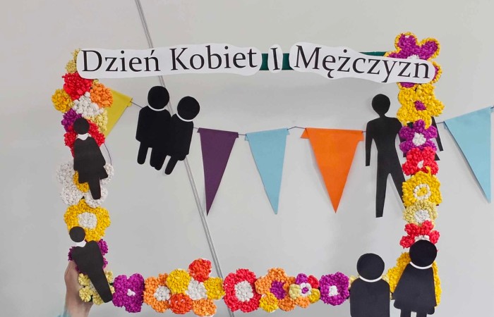 Dekoracyjna ramka z napisem „Dzień Kobiet i Mężczyzn”, ozdobiona kolorowymi kwiatami z bibuły i sylwetkami postaci, trzymana przez osobę w niebieskim swetrze. W tle kolorowe girlandy zawieszone na ścianie.