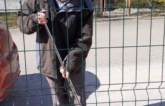 Starszy pan zamiata piasek z chodnika miotłą o srebrnym kiju. Jest ciepło ubrany w czarne ciuchy.