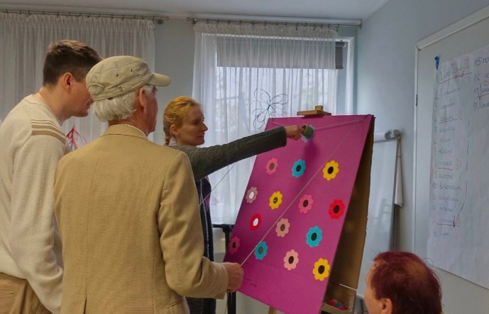 Grupa osób bierze udział w grze zręcznościowej przy tablicy z kolorowymi kwiatkami. Kobieta w szarym swetrze wskazuje na planszę, starszy mężczyzna w beżowej marynarce trzyma sznurek, a obok nich stoi młodszy mężczyzna w białym swetrze. Na krześle siedzi kobieta z krótkimi czerwonymi włosami, uśmiechając się i obserwując grę.