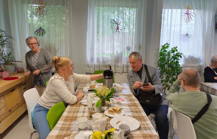 Spotkanie przy stole – kobieta w białym swetrze nalewa kawę z termosu dla mężczyzny w szarym garniturze, który trzyma telefon i rozmawia z innymi uczestnikami. Na stole filiżanki, talerzyki z ciastem i żółte kwiaty w wazonie. W tle inne osoby oraz duże okna z ozdobami.