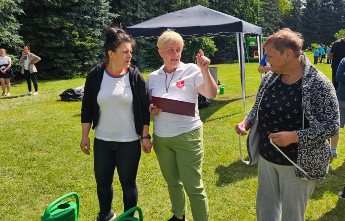 Trzy kobiety stoją na trawie, jedna z nich, sędzia, tłumaczy im zasady konkurencji sportowej, przed nimi stoją dwie zielone konewki które są elementem konkurencji. W tle widać namiot i innych ludzi uczestniczących w zawodach. Świeci słońce i jest ładna pogoda 