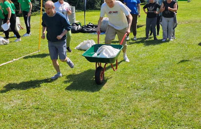 Konkurencja sportowa w której dwóch mężczyzn biegnie wzdłuż linii po trawie, jeden z nich pcha zielono czerwoną taczkę z workiem. W tle widać sędzinę odmierzającą czas w jakim uda im się zaliczy konkurencję, innych członków drużyny którzy dopingują zawodników oraz przygotowujący się drugi zespół